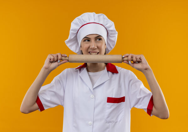 厨师穿着厨师制服的年轻恼怒的白人厨师女孩拿着并假装咬着橘黄色背景上的擀面杖 还有复制空间卷制服假装