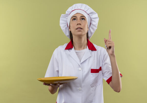 厨师穿着厨师制服的年轻白人厨师女孩拿着盘子 孤立地指着绿色背景和复印空间惊喜点制服