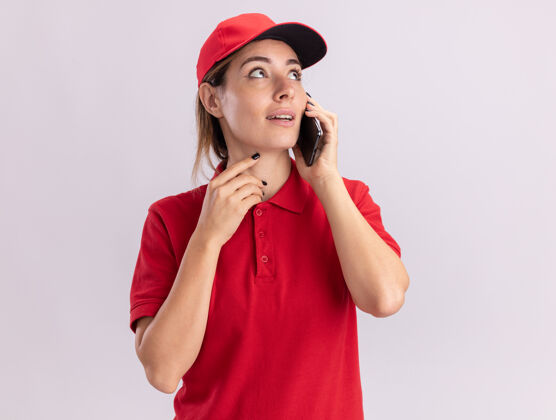 衣服印象深刻的年轻漂亮的女送货员穿着制服在电话里交谈 看着隔离在白墙上的一面电话制服公民