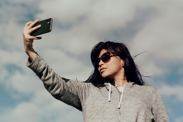 年轻戴着墨镜的年轻女子在多云的蓝天下用手机拍照漂亮自拍肖像