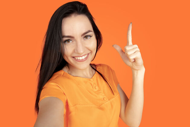 行政橙色工作室背景上的白种人年轻女子肖像穿着衬衫的漂亮黑发女性模特人类情感的概念 面部表情 销售 广告广告空间指向 展示 微笑人类霓虹灯人
