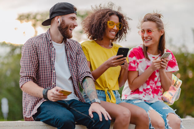 享受快乐的年轻朋友们坐在公园里用智能手机微笑 男人和女人玩得开心友谊团体耳机