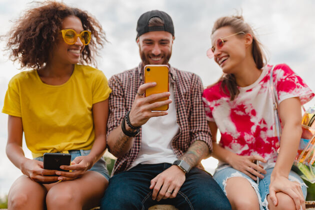 积极快乐的年轻朋友坐在公园里用智能手机微笑公司欢笑青春年轻