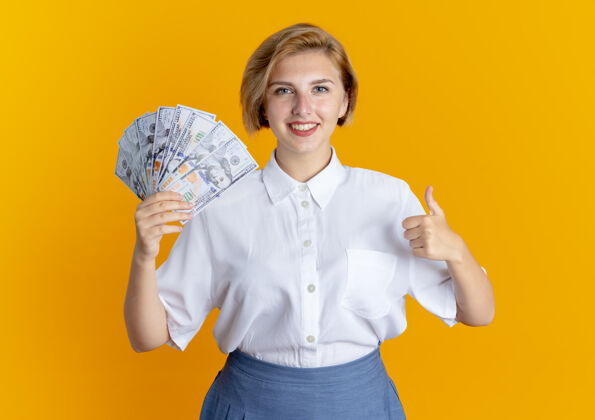 拇指年轻微笑的金发俄罗斯女孩拿着钱 竖起大拇指孤立在橙色背景与复制空间抱着金发微笑