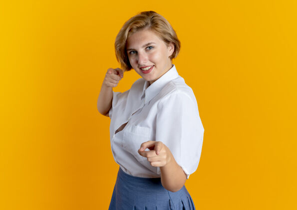 侧身年轻的微笑的金发俄罗斯女孩站在一旁指着相机孤立的橙色背景与复制空间镜头指着年轻人