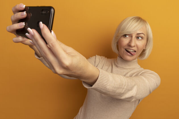表情快乐美丽的金发斯拉夫女人结结巴巴的舌头 看着手机自拍隔离在橙色的墙上姿势卡住自拍