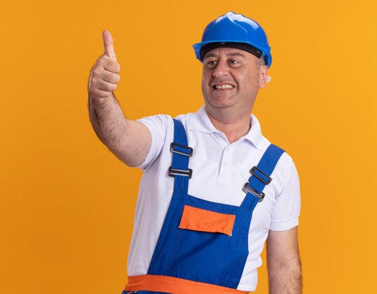 人微笑的成年建筑工人身着制服 竖起大拇指孤立地站在橙色的墙上人人建筑者