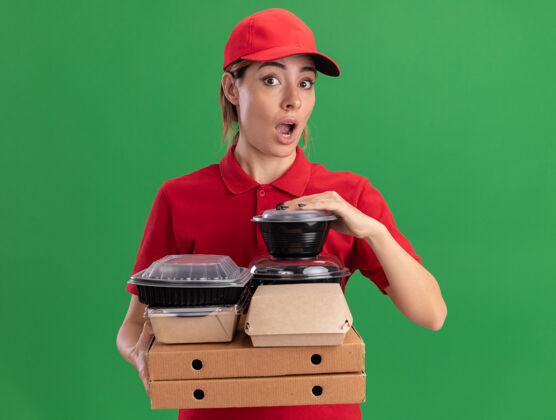 脸令人印象深刻的年轻漂亮的女送货员穿着制服拿着纸食品包装和比萨饼盒上的容器隔离在绿色的墙壁上盒子制服站着