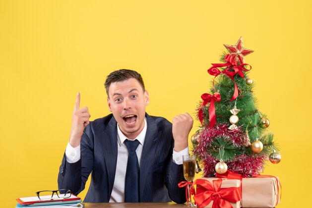 附近正面图惊奇的男人手指着坐在圣诞树旁的桌子和黄色背景上的礼物树行政人员惊奇的男人