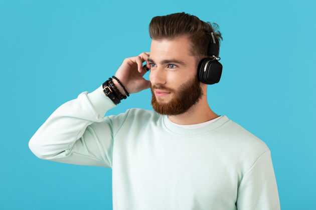 扬声器时尚迷人的年轻胡须男子在无线耳机上听音乐现代风格自信的心情隔离在蓝色的墙上现代时髦乐趣