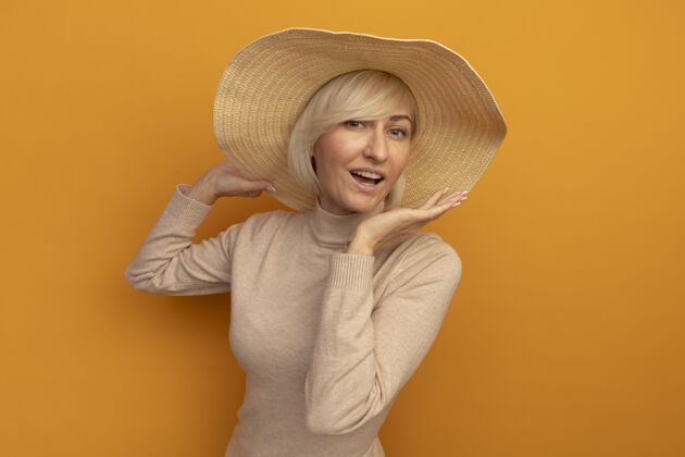漂亮快乐美丽的金发斯拉夫女人 戴着沙滩帽 手张开 孤立地站在橙色的墙上开放感觉帽子
