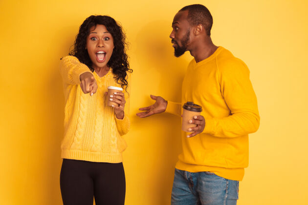 情感年轻感性的非裔美国人男女 穿着黄色背景的明亮休闲服美丽的情侣人类情感的概念 面部表情 关系 广告喝咖啡和指点指向年轻姿势