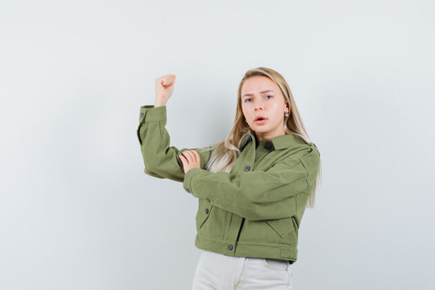 成人穿着绿色夹克的年轻女性 牛仔裤检查手臂肌肉 看起来很专注 正面视图女孩年轻焦点