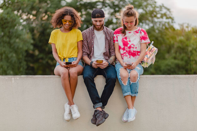 多样性快乐的年轻朋友坐在公园里用智能手机微笑公司时髦女人手机