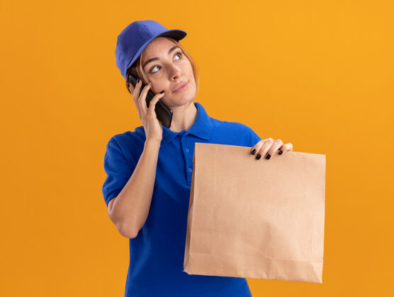 站穿着制服的年轻漂亮的女送货员拿着纸包 看着橘色墙上孤立的一面讲电话女孩制服衣服