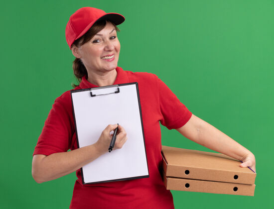 女人身穿红色制服 头戴鸭舌帽的中年女送货员拿着披萨盒和剪贴板 手里拿着空白页和钢笔 微笑着看着前面 站在绿色的墙上要求签名盒子空白帽子