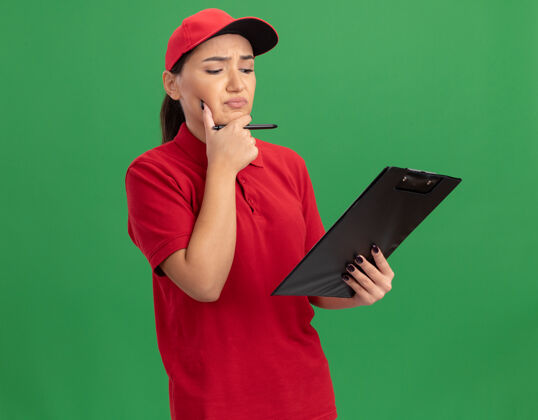 帽子身穿红色制服 戴着便笺和铅笔帽的年轻女送货员 脸上带着沉思的表情 站在绿色的墙上思考立场思考脸