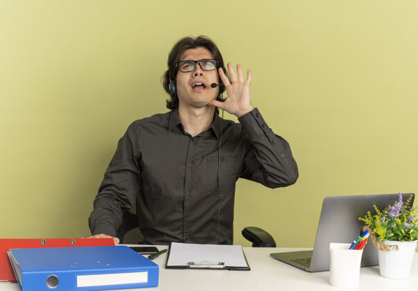 耳机戴着耳机坐在办公桌旁 拿着办公工具 用笔记本电脑 假装在绿色背景下给一个被隔离的人打电话 留着复印空间办公室年轻工人