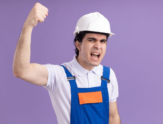 拳头身穿施工制服 头戴安全帽的年轻建筑工人站在紫色的墙上 怒气冲冲地高喊着脸制服举起