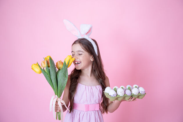 母亲节快乐一个有复活节兔子耳朵的小女孩 手里拿着一盘鸡蛋 在粉红色的墙上嗅着一束郁金香复活节彩蛋兔子孩子