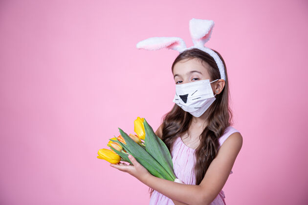 节日一个戴着医用口罩 戴着复活节兔子耳朵的小女孩手里拿着一束郁金香放在粉红色的墙上小花兔子