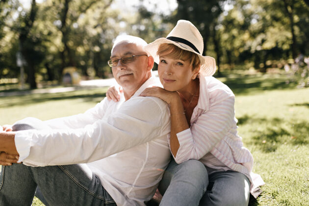 姿势漂亮的女人 金发 戴着时髦的帽子 穿着粉色的衬衫 坐在草地上 男人留着胡子 穿着白色的衣服 在公园里祖父母青年情感