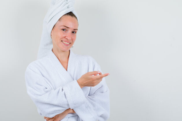 干净年轻女士指着右边穿着白色浴衣 毛巾 看上去很快乐正视图人护理头发