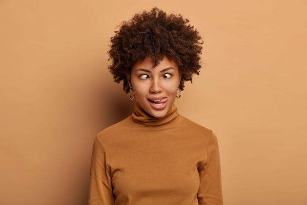 高领毛衣疯狂的美国黑人女人做滑稽的脸 伸出舌头和交叉的眼睛 傻瓜周围 穿着休闲的棕色高领毛衣情绪漫画笑话
