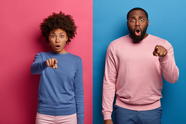 朋友情绪激动的美国黑人妇女和男人用食指指着你穿着五颜六色的衣服对一些可怕的事情做出反应 站在工作室里对着粉色毛衣黑发拼图