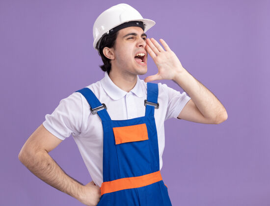头盔身穿施工制服 戴安全帽的年轻建筑工人站在紫色的墙上 用手在嘴边喊或叫人呼喊制服呼叫