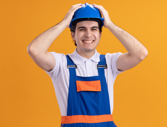 快乐年轻的建筑工人身穿建筑制服 戴着安全帽 手持铁锤 双手放在头上 站在橙色的墙上 高兴而兴奋地看着前方兴奋建设者锤子