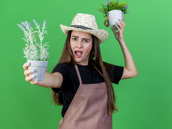 穿愤怒美丽的园丁女孩穿着制服 戴着园艺帽 举着花盆里的鲜花 在绿色的背景下与世隔绝举行女孩提高