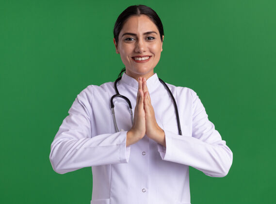 手掌年轻的女医生 穿着医用外套 手持听诊器 双手合十 站在绿色的墙上 微笑着看着前方微笑握着脸