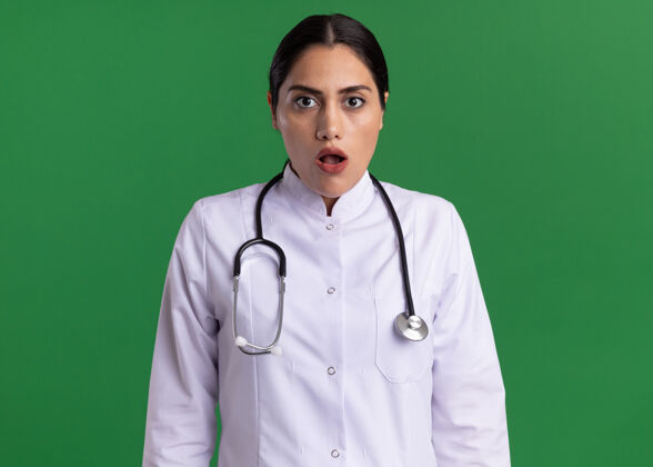 医生年轻的女医生 穿着医用外套 脖子上戴着听诊器 站在绿色的墙上 困惑而担心地看着前方脖子外套年轻
