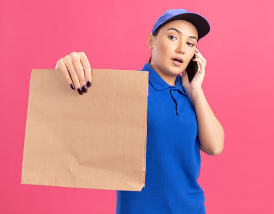 脸身穿蓝色制服 头戴鸭舌帽的年轻送货员站在粉色的墙上 严肃地看着包裹 一边讲着手机包装拿着帽子