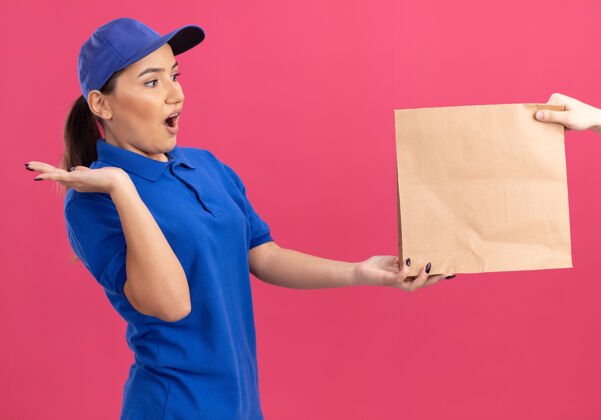 递送身穿蓝色制服 戴着帽子的年轻送货员站在粉色的墙上 一边接受纸包裹 一边兴奋不已年轻制服感情