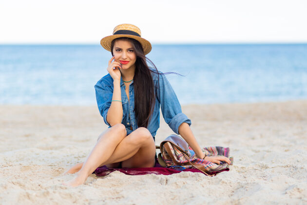 享受美丽的女人 棕褐色的身体 丰满的红唇和长长的腿 在热带阳光明媚的海滩上摆姿势穿着短上衣 短裤和草帽沙子放松垫子