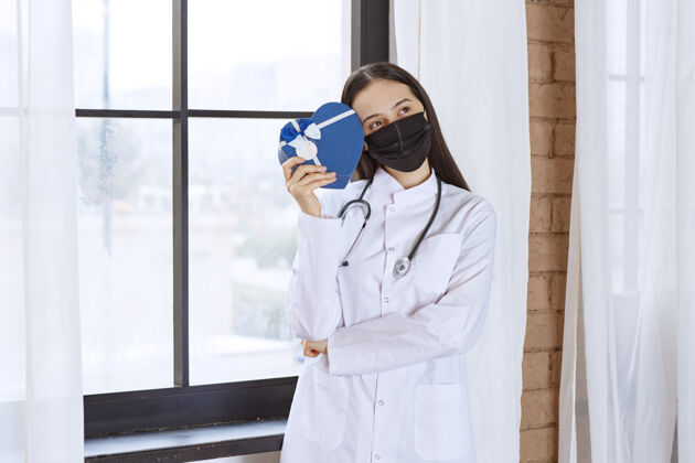 阳性医生拿着听诊器和黑色口罩 手里拿着一个蓝色心形礼盒 看上去既困惑又不确定模特制服健康