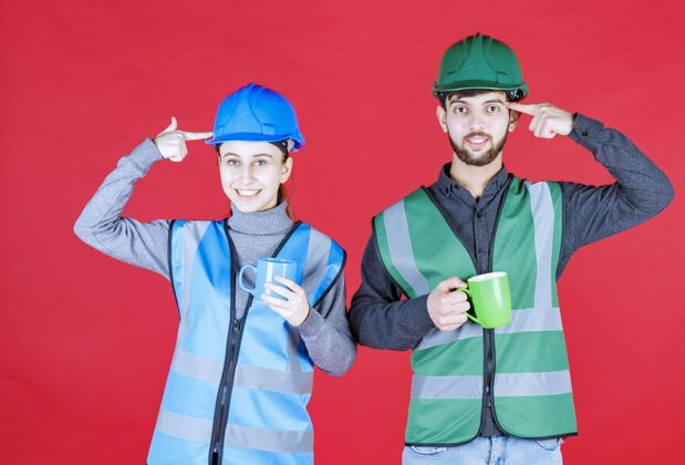 工人男女工程师戴着头盔 拿着蓝色和绿色的杯子 思考着新的想法休息水化制服