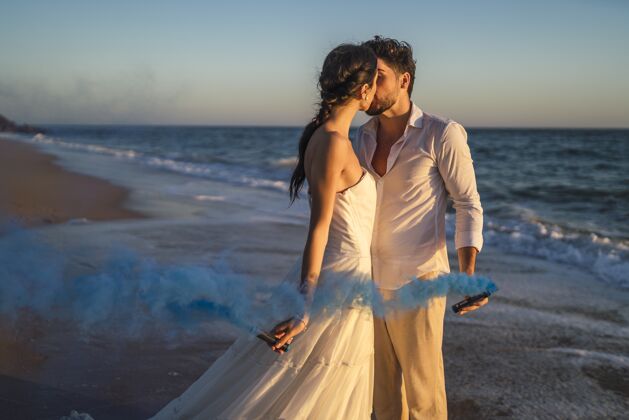 浪漫一对高加索情侣在婚礼上手持蓝色的烟在沙滩上亲吻背景人夫妻