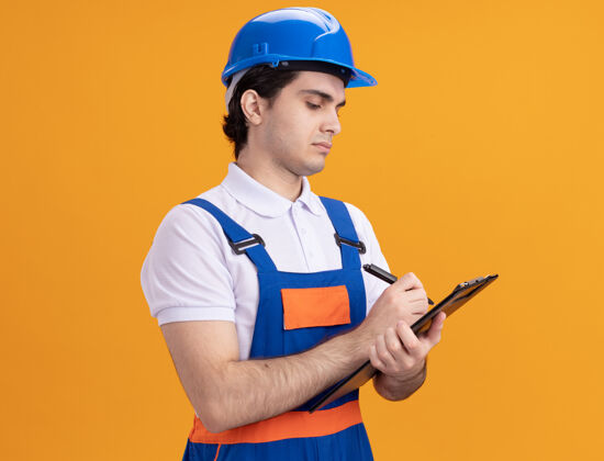 建筑工人年轻的建筑工人穿着建筑制服 戴着安全帽 手里拿着写字板 手里拿着钢笔 自信地站在橙色的墙上拿着头盔年轻人