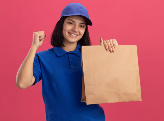 女孩身穿蓝色制服 头戴鸭舌帽的年轻送货员手持纸包握紧拳头站在粉色的墙上 快乐而兴奋制服拳头年轻