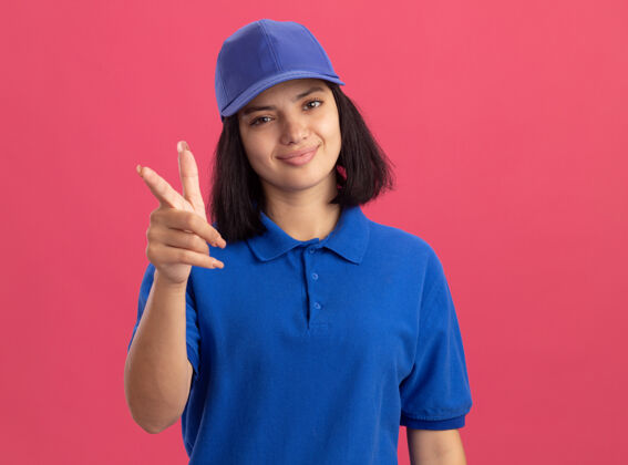 交货身穿蓝色制服 戴着帽子 食指友好地站在粉色墙壁上微笑的年轻送货女孩朋友手指微笑