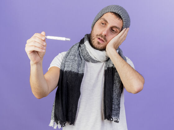 关心一个戴着冬天帽子 戴着围巾 手放在脸颊上 隔离在紫色背景上 看着温度计的年轻病人体温计帽子戴