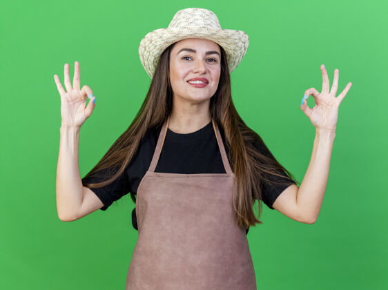 手势微笑美丽的园丁女孩穿着制服 戴着园艺帽 在绿色的背景上显示出很好的手势穿制服园艺