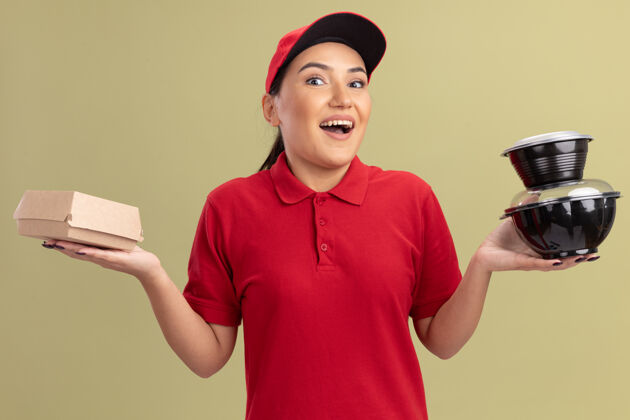 拿着身着红色制服 头戴鸭舌帽 面带微笑地站在绿色墙壁上 拿着食品包的快乐的年轻送货员望着前方递送脸盒子