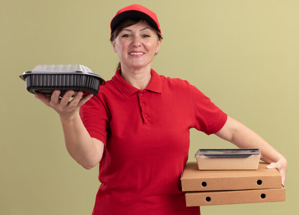 女人身穿红色制服 头戴鸭舌帽的中年女送货员拿着披萨盒和食品包 微笑着自信地站在绿色的墙上看着前方披萨送货微笑