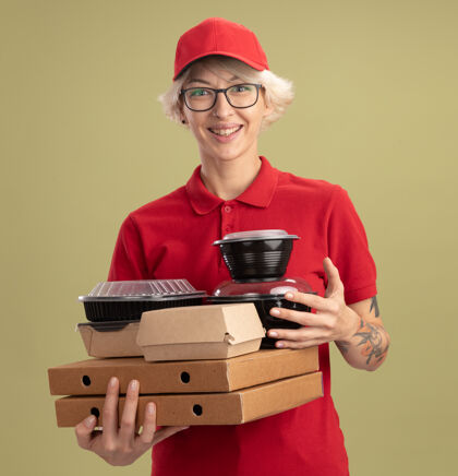 站着身着红色制服 戴着眼镜 拿着比萨饼盒和食品包的快乐的年轻送货员站在绿色的墙上开心地微笑着帽子欢呼女人