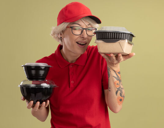 帽子身着红色制服 戴着眼镜 手里拿着食品包 站在绿色墙壁上高兴地微笑着的年轻送货员送货抱着眼镜