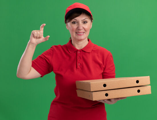 制服身穿红色制服 头戴鸭舌帽的中年送货妇女拿着披萨盒 展示着小施的手势 手指看着前面 站在绿色的墙上 开心地微笑着站着送货拿着